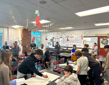 Students are actively engaged in various activities inside a classroom decorated with educational posters, including a large periodic table. There are hanging decorations from the ceiling, and the room is lively with students working at tables, some standing and others seated.