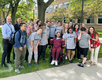 Group of students and teachers posing outdoors on a sunny day, with trees and a school building in the background. Some students are wearing shirts with the