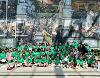 Group of children wearing green shirts seated in front of the Detroit Industry Murals by Diego Rivera at the Detroit Institute of Arts.