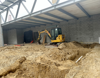terior of a construction site with a Caterpillar excavator, piles of dirt, and partial brick walls under a metal roof.
