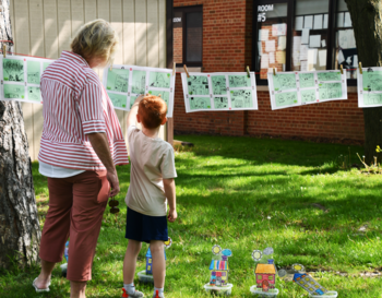 An adult and a child viewing artwork displayed on a line between trees at a school event.