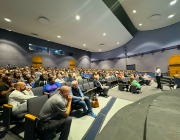 Audience seated in a large auditorium listening to a speaker standing on stage.