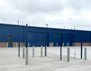 A large blue warehouse with multiple bays and a concrete pavement in front, under a cloudy sky.