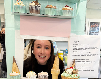 A smiling person peeking through a display window surrounded by various desserts including cupcakes and ice cream cones