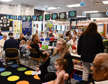 Classroom filled with adults and children gathered for an event, featuring colorful decorations and educational posters on the walls.