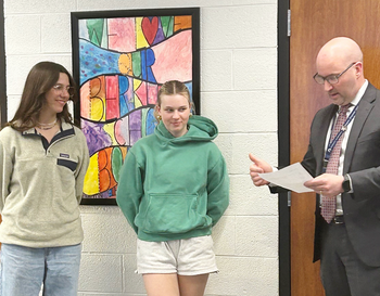 Two students stand beside a colorful abstract stained artwork, listening as an educator holding a paper explains something, in a school setting.