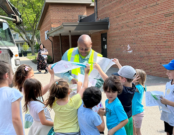 A waste management worker in a reflective vest showing a map to a group of enthusiastic children outside a brick building.