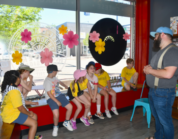A group of children in yellow shirts sitting inside of a store, attentively listening to an adult speaker. The shop has a colorful decor with large flower decals on the windows.