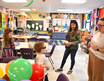 Interior view of a vibrant classroom with colorful decorations and several large balloons. A child and two adults they engaged in an interactive activity.