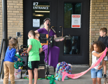 A musician playing a guitar and singing into a microphone at a school entrance labeled