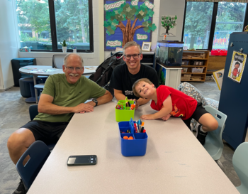 Three people smiling at a table in a classroom, with art supplies in front of them, under a bulletin board decorated with a tree and apples.