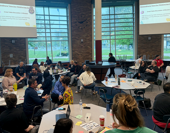 Attendees seated at tables during a workshop in a spacious room with large windows overlooking a grassy area, with projection screens displaying educational material about microcredentials and student success strategies.