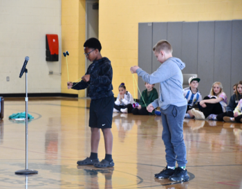 Two students participate in a talent show in a school gymnasium, doing yo-yo tricks as an audience of peers watches from the background.