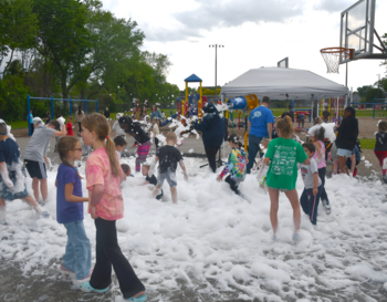 Children playing in foam at a community outdoor event near a basketball court, with playground equipment visible in the background.