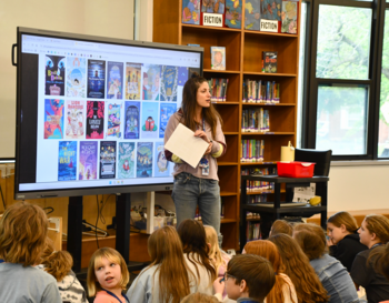 A person stands in front of a large screen displaying book covers during a presentation to a group of attentive children in a library.