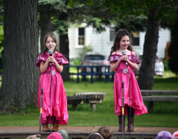 Two children in matching pink dresses performing on an outdoor stage, holding microphones.