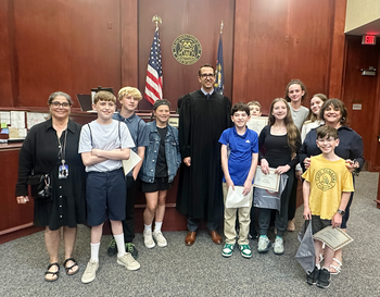 Group of people, including a judge in a robe, posing in a courtroom near the American and state flags.