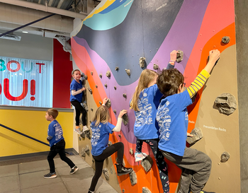 Children in blue shirts climbing on an indoor rock wall.