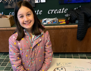 A child standing in front of a display, proudly presenting a colorful artwork captioned 