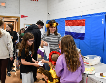 Group of people, including children and adults, interacting at a cultural event booth draped with the flag of the Netherlands. Some individuals are looking at materials on the table while others are engaged in conversation.