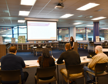 A person presenting to a small group in a media center, with audience members seated at tables. A projection screen displays a presentation titled