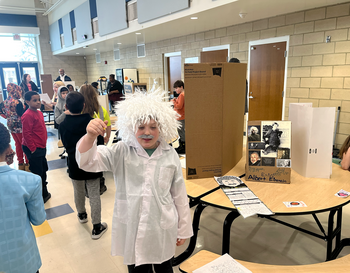 A child dressed as Albert Einstein with wild white hair, giving a thumbs up in a crowded school hall during a presentation event. There are displays and other children around the room.