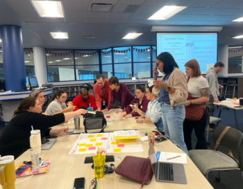 Group of individuals collaboratively working around a table with laptops and papers, engaged in a workshop inside a room with a whiteboard that includes notes and a projection screen displaying a presentation.