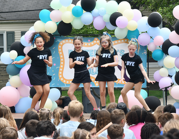 Four children perform a dance on stage at a school event, wearing matching black outfits with the word