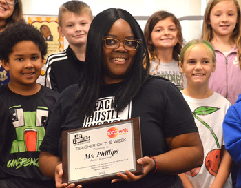 Angell Elementary School's music teacher, Ashley Phillips receiving a plaque for WNIC teacher of the week with students smiling in the background