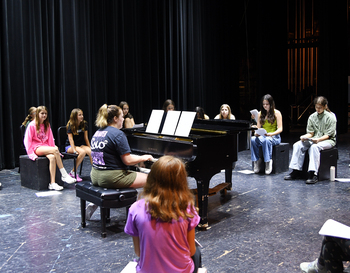 Ten students sit on blocks around a piano listening to an instructor.