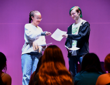 Two people are standing on a stage, exchanging papers in front of an audience with a purple background.