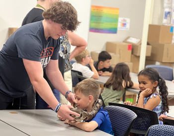 A person assisting a young child with an activity at a table in a classroom setting where a snake is being placed around the students shoulders with other children seated around.