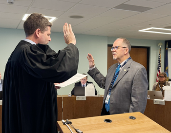 A person in judicial robes administering an oath to another person in a business suit, standing in a meeting room with other seated individuals in the background.