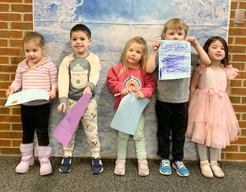 Five children stand in front of a winter-themed backdrop, each holding a piece of artwork.