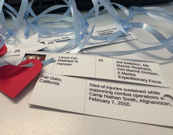 A memorial card for Lance Cpl. Matthias N. Hanson with ribbons and a heart-shaped pendant on a desk. The card indicates he was from Fair Oaks, California, belonging to the 3rd Battalion, 6th Marine Regiment, 2nd Marine Division, II Marine Expeditionary Force and passed away on February 7, 2010, in Afghanistan.