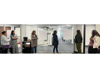 Six individuals standing in a modern office space, engaging in a discussion near whiteboards covered with notes and markers.