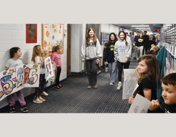 Children and teenagers in a school hallway holding colorful handmade signs, with some wearing Michigan State apparel.