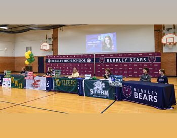 Signing event at Berkley High School gymnasium with students seated at tables, each featuring a banner from various universities.