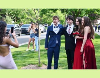 A group of teenagers in formal attire posing for a photo at a prom event, with another person taking their picture.