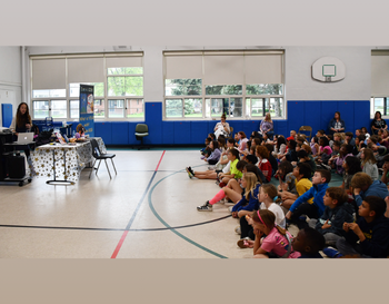 A group of students sitting on the floor in a school gymnasium, attentively listening to a speaker at a podium.