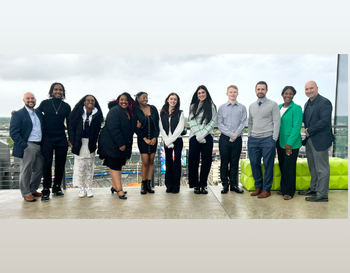 Group of students and educational professionals posing together dressed in business casual attire at an outdoor event, with a cloudy sky in the background.