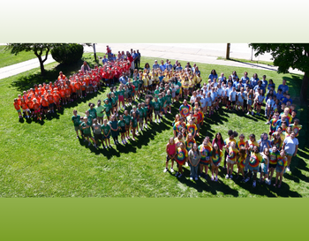 Large group of individuals wearing color-coordinated t-shirts arranged in a shape of the number 100 in a sunny outdoor setting.