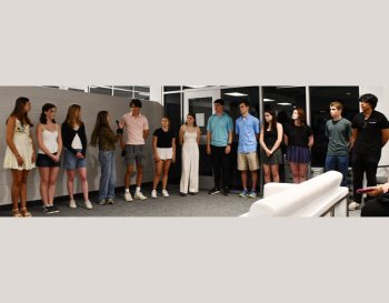 A group of teenagers standing in a row inside a room, dressed in casual and formal wear, engaged in a presentation or event.