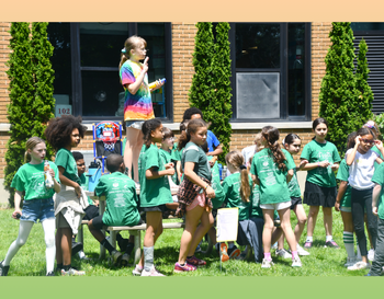 A group of children wearing green T-shirts and one child standing on a bench speaking into a microphone during an outdoor school event.