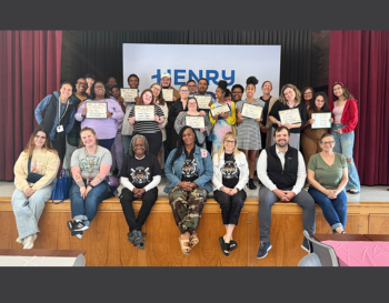 Group of people posing with certificates in front of a banner where the words 'Henry' can be seen behind people on a stage.