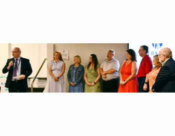 A group of eight individuals standing during a presentation at an event, with one person speaking into a microphone.