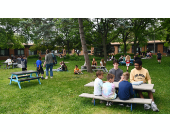 Students and teachers enjoying outdoor activities on the grassy lawn of a school campus, with some seated at picnic tables and others standing or sitting in groups.