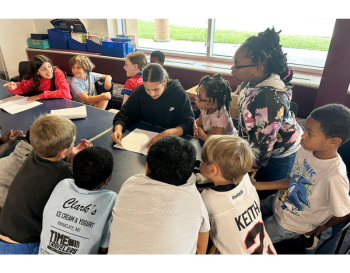 Group of children gathered around a table in a classroom, with some wearing sports jerseys, actively engaged in a discussion led by one person holding an open book.