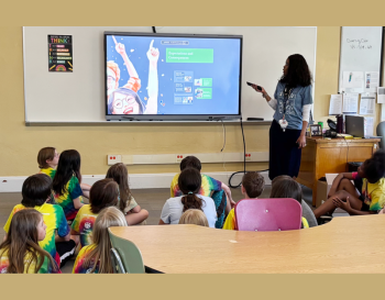 Teacher presenting to a group of students wearing matching t-shirts in a classroom. The presentation slide on the screen features diagrams.