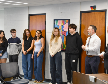 Group of six students and two teachers standing in a classroom, interacting near a whiteboard with colorful artwork in the background.